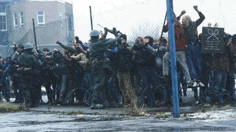 A still from the films showing people contained within tennis courts, clamouring at the wire fencing, and wardens with hard hats and guns trying to keep them contained.