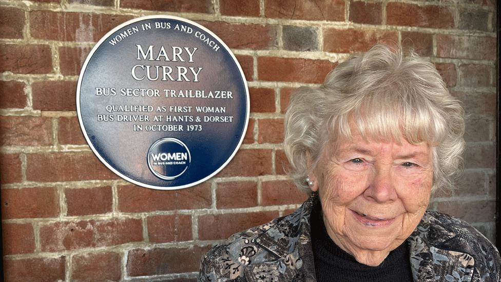 Mary Curry standing next to the blue plaque dedicated to her. It says: "Mary Curry. Bus sector trailblazer. Qualified as first woman bus driver at Hants & Dorset in October 1973." She has short grey hair, and is wearing a grey patterned jacket over a black top.