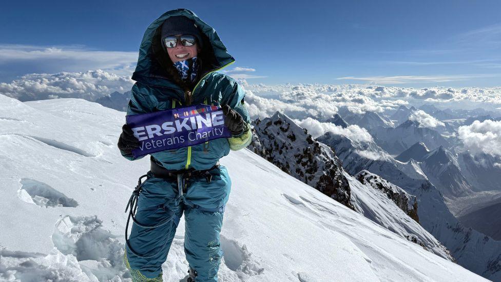 Kirsty Mack is smiling at the camera. She is wearing climbing gear and sunglasses at the top of a mountain, with blue skies and snowy peaks behind her.