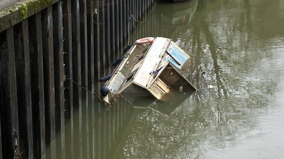 A boat on it's side slowly sinking in the river with a taut rope leaving it hanging from the side