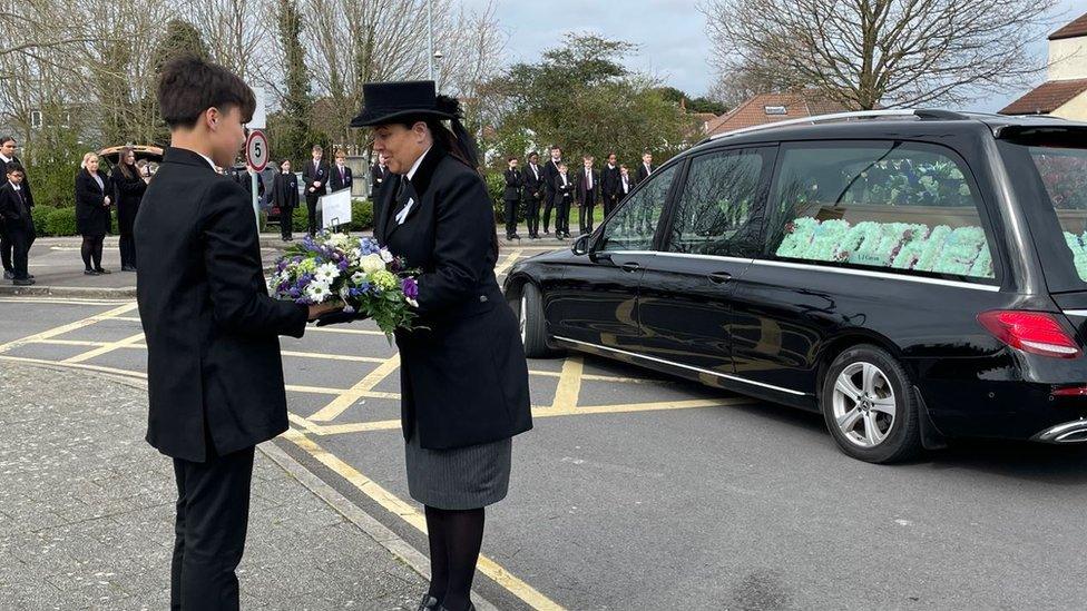 A teenage boy dressed in a black suit hands a bunch of flowers to the funeral director whilst the car carrying Max's coffin is seen in the background