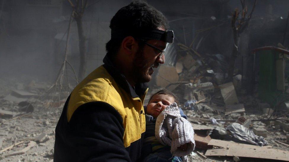 Amid rubble and smoke, a man wearing a headlamp and yellow jacket carries a baby in distress