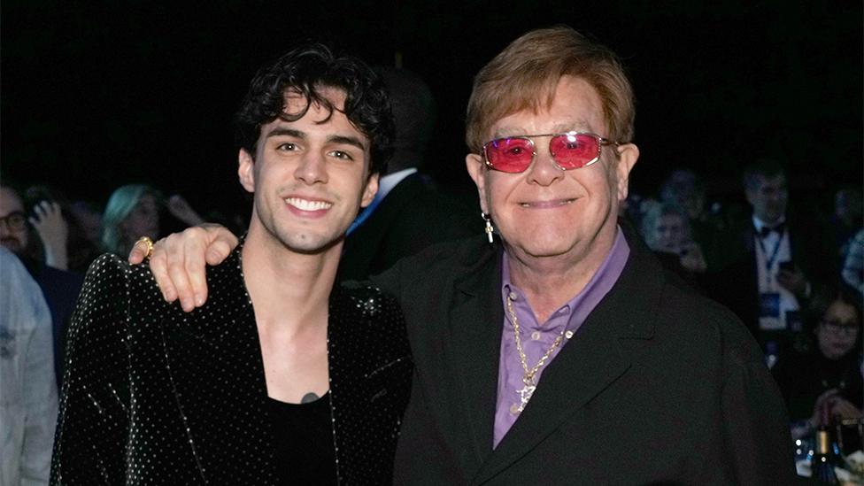 Stephen Sanchez on the left and Elton John on the right looking at the camera, with Elton having his arm around Stephen's right shoulder. Stephen is wearing a black blazer with black vest, while Elton has a black blazer with a purple shirt and red tinted glasses.