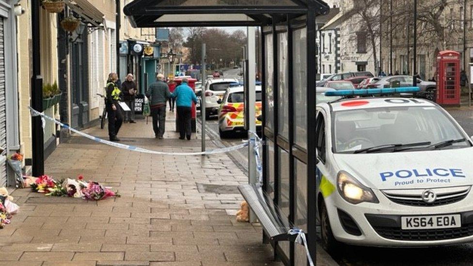 A police car and cordon at the scene, along with floral tributes laid on the ground