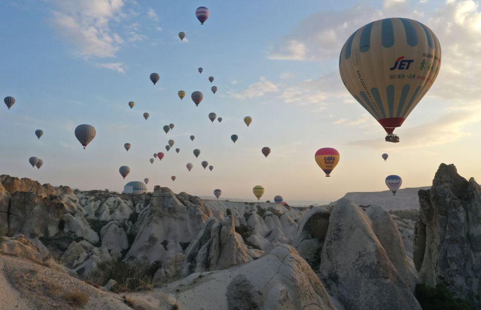 Hot air balloons fly over Cappadocia, a UNESCO World Heritage Site.  on October 14, 2024.  Cone-shaped rock formations are in the foreground.  Over twenty balloons of various colour, rise up at various distances into a pale blue sky.  Faint clouds are on the right hand side.