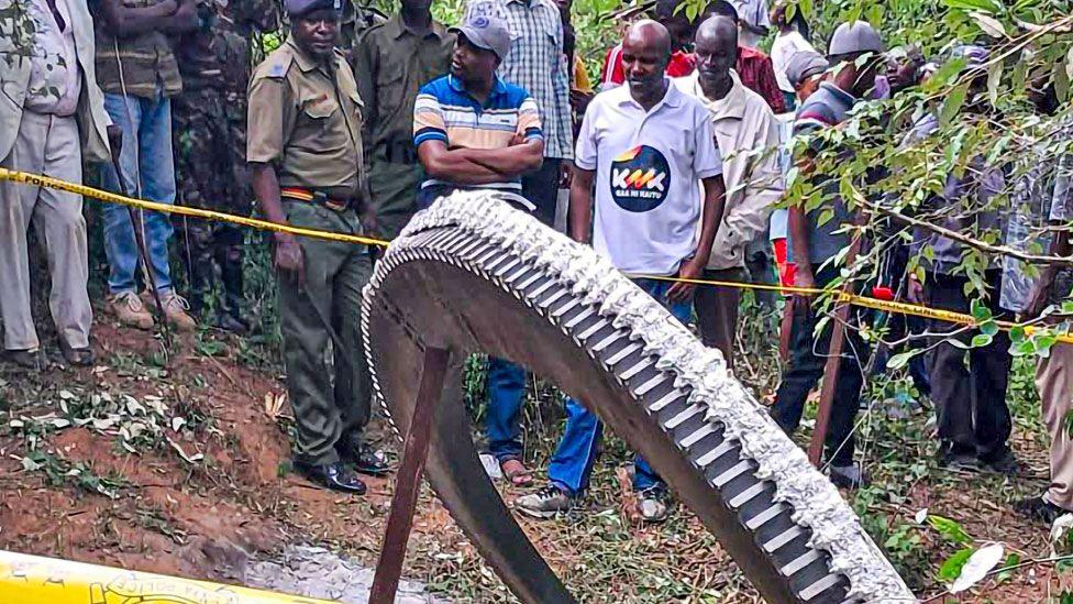 A crowd, including police officers, stand behind yellow tape looking on at a giant metal ring which fell from space on to farmland in Mukuku, Kenya 