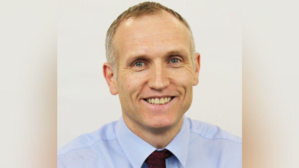 A headshot of a man who is smiling into the camera. He is wearing a blue shirt and burgundy tie that is slightly off centre.