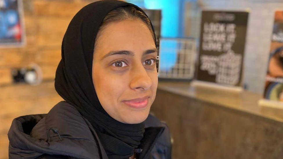 Mariam smiles as she stands in the cafe in a black headscarf and coat.