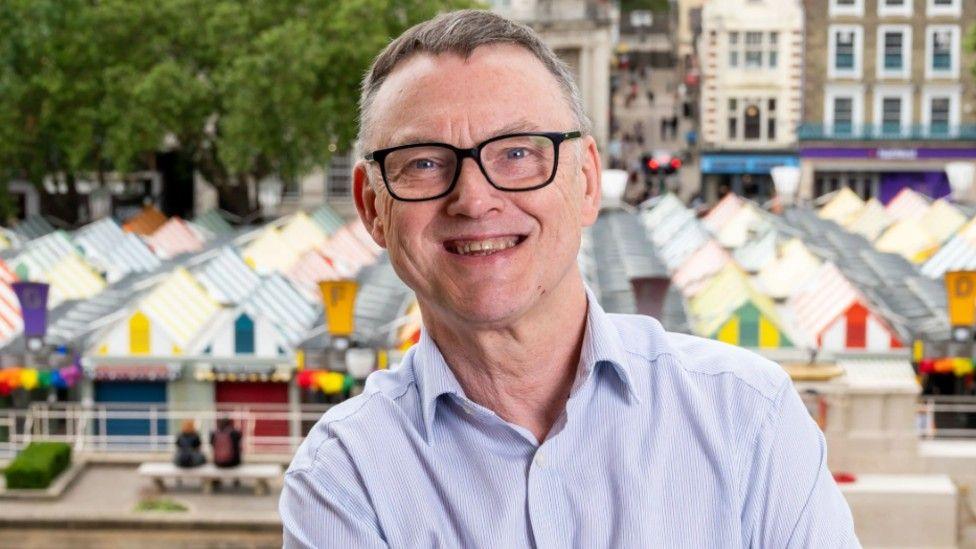 Mike Stonard, a man in a blue stiped shirt. He is looking at the camera and smiling. He is wearing black rimmed glasses and is stood at the top of Norwich Market.