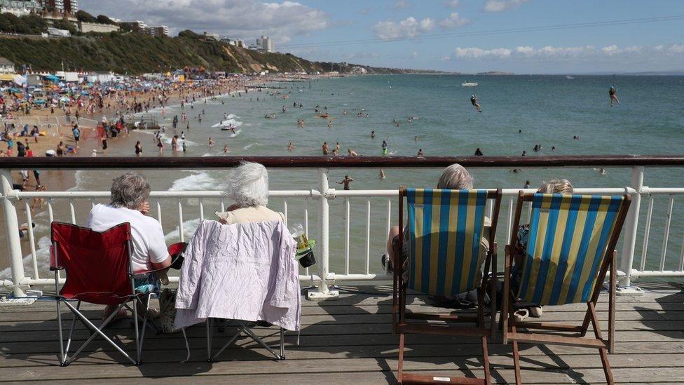 Rear view of four white-haired people sitting on deckchairs and camping chairs on Bournemouth pier, looking along the beach and shoreline which is filled with people.