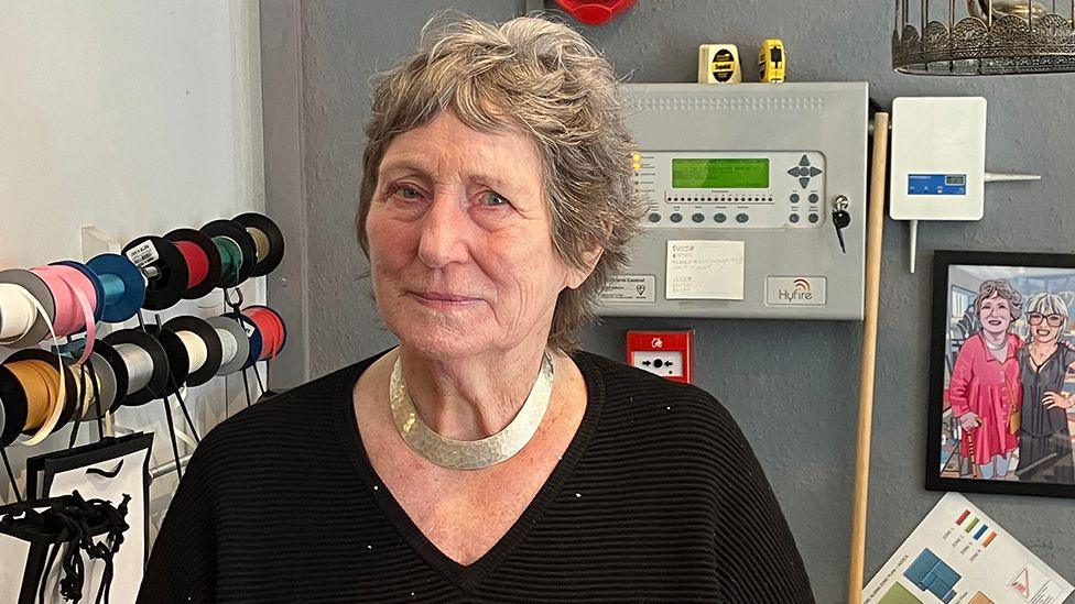 Head and shoulders shot of Maureen with short grey hair wearing a black jumper standing behind the till of her gift shop