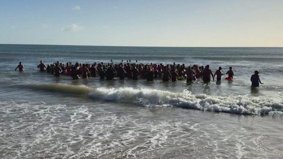 Dozens of people in a group in the sea with small waves crashing onto the beach.