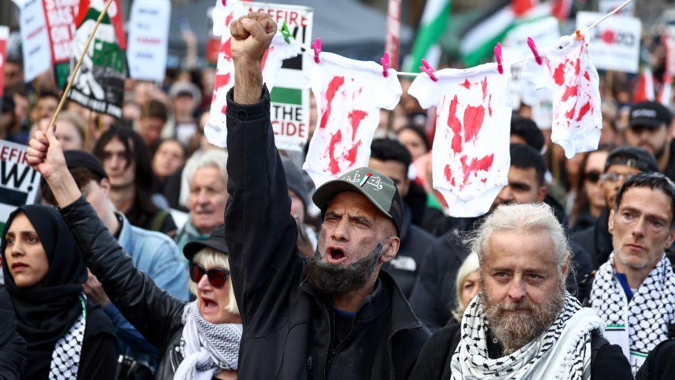 Protesters raised their arms in the air. Several are carrying signs with white red and green colours, while some hold up white children's clothes stained with red. 