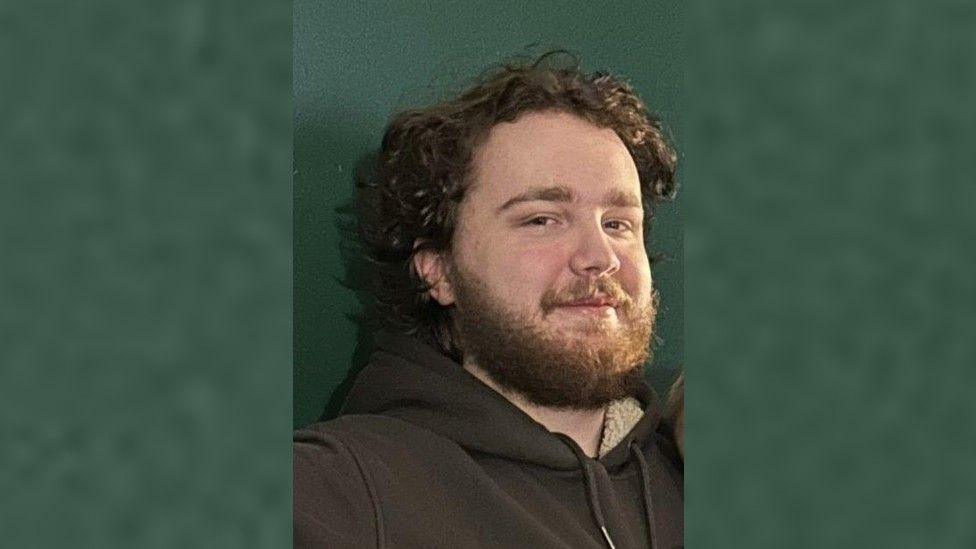 Man with curly brown hair and brown beard, smiling and looking at the camera with a dark green wall in the background