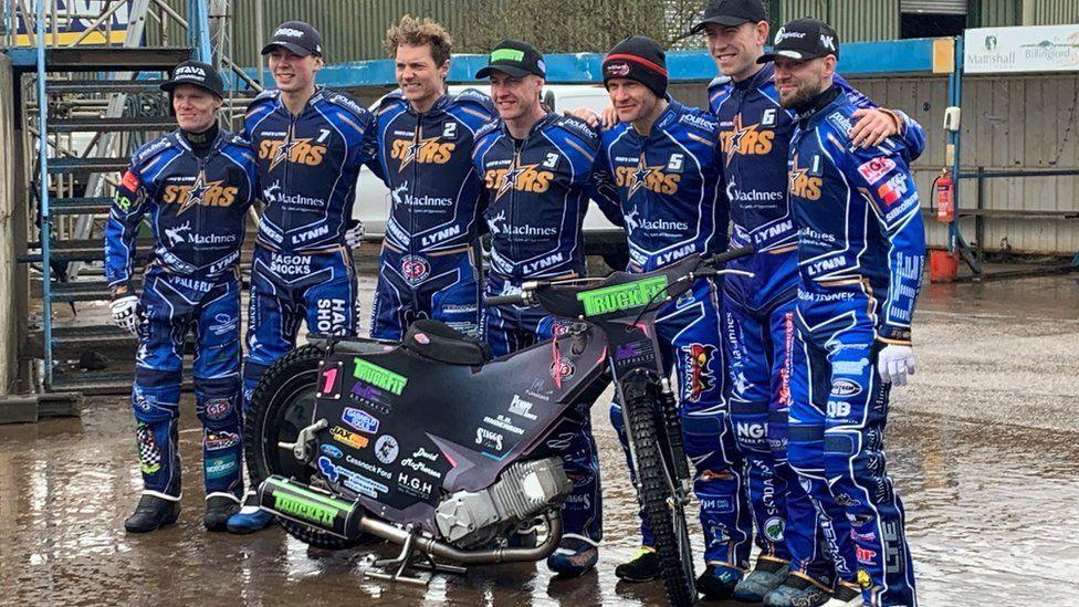 Eight men wearing blue King's Lynn Stars leathers standing around a motorbike