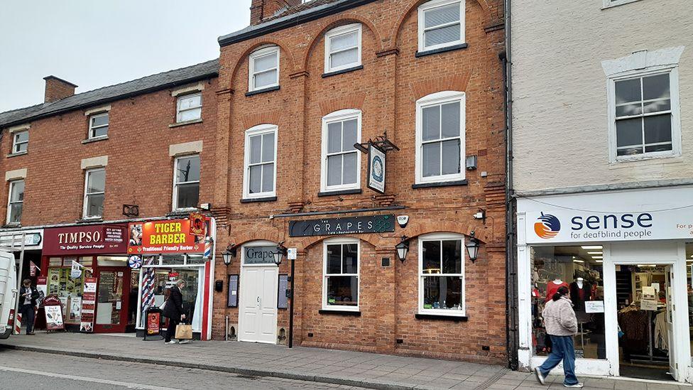 The outside of the Grapes in Sleaford which is a narrow, three-storey brick building located between a row of shops