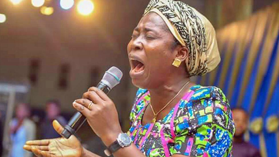 Osinachi Nwachukwu with her eyes closed as she sings into a microphone. She is wearing a cream headwrap with a black pattern, earrings and a necklace and a blue, black green and pink patterned dress.