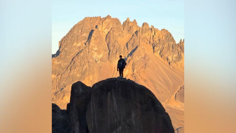 A silhouette of Aaron on top of a mountain against another mountain in the distance and a blue sky