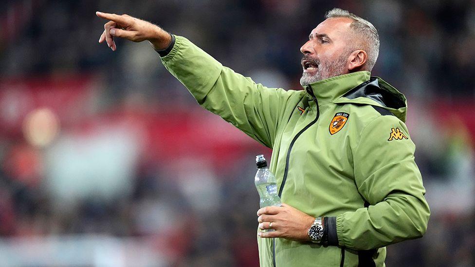 Hull City manager Tim Walter gesticulating form the touchline wearing a lime green jacket and holding a bottle of water