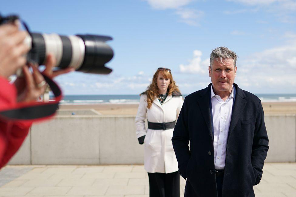 Labour Party leader Sir Keir Starmer and Angela Rayner, deputy leader and chairwoman of the Labour Party visit Seaton Carew seafront on 1 May 2021 in Seaton Carew. Sir Keir is in the foreground with his hands in his pockets, with Rayner behind him. A camera shoots a photo, with the beach behind them.