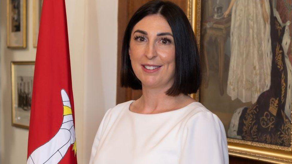 A female politician with black hair parted in the middle, wearing a white top. She is standing in front of an Isle of Man flag on a standing pole and an old political portrait in a government office. 