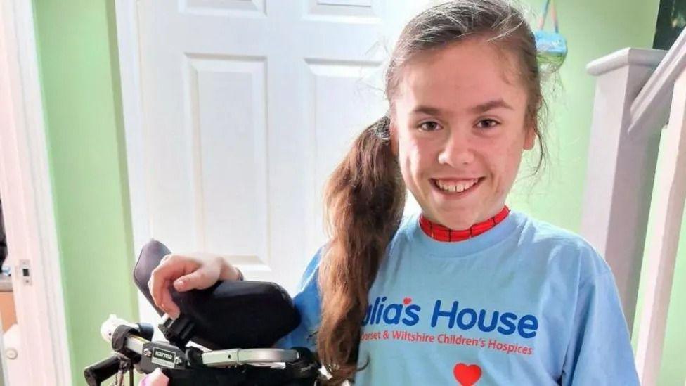 Carmela, from Wareham wearing a light blue julia's house t-shirt over her superhero costume and stood next to her wheelchair