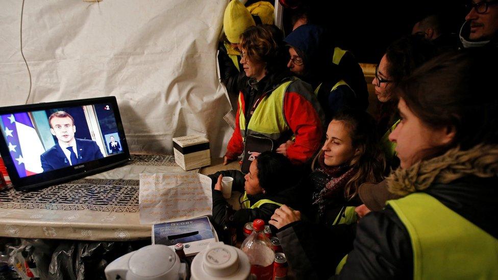 Protesters wearing yellow vests watch French President Emmanuel Macron on a TV screen