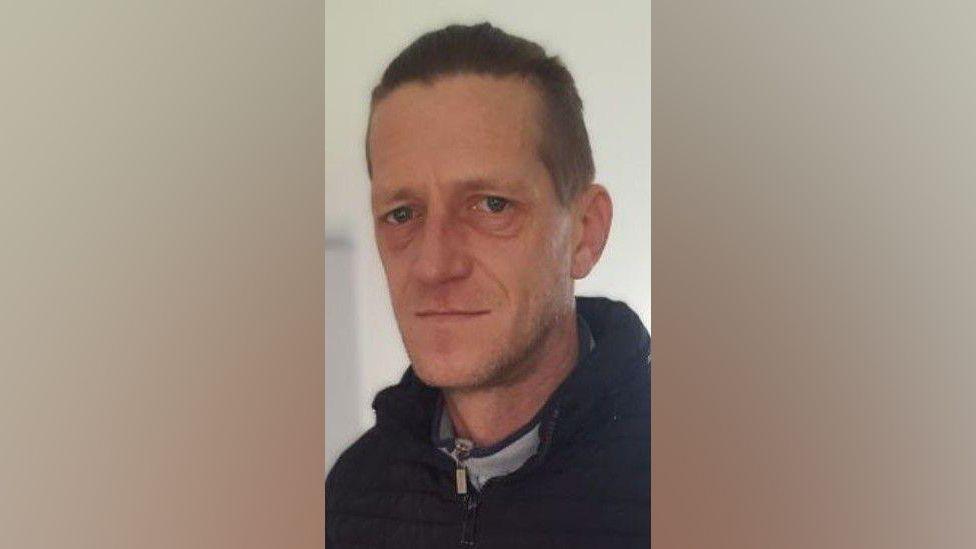 Head and shoulders shot of Vadims Zmicerevskis who has short hair and is wearing a black zipped up jacket and coat. There is a beige wall in the background.