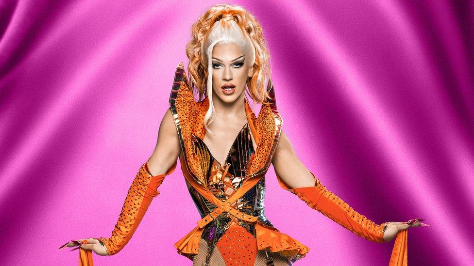 Drag queen Alexis Saint Pete standing in front of a purple background with his arms outstretched. He's got blonde hair tied up with orange highlights at the end and is wearing orange elbow length gloves. He's also wearing an orange, metallic waistcoat style bodice with fishnet stockings on.