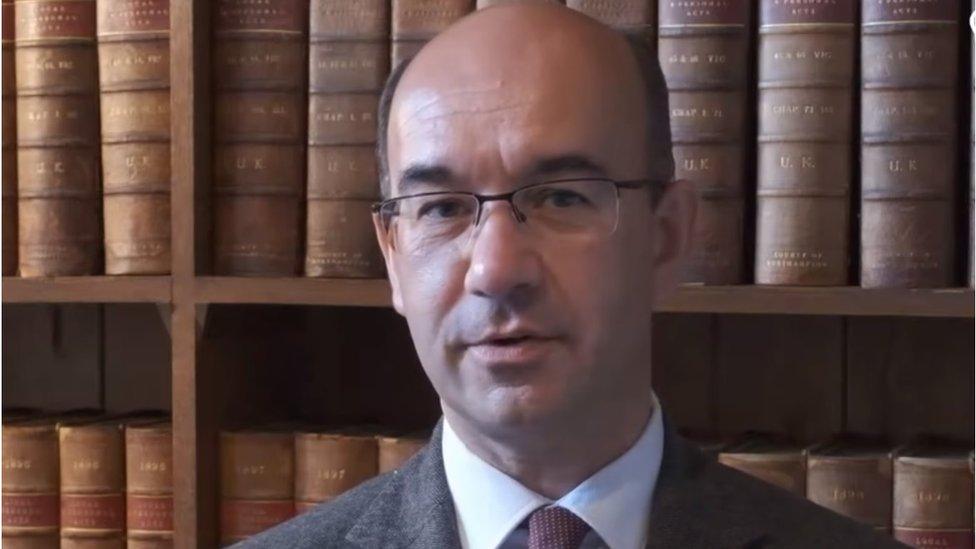 Man with short dark hair and glasses stands in front of leather-bound books