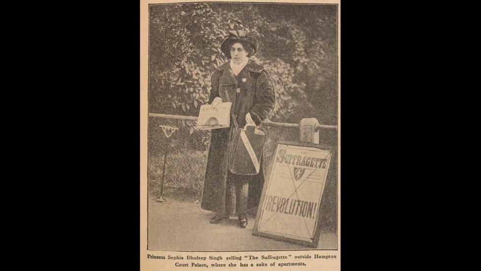 Sophia seen in a heavy coat and a hat holding newspapers and standing next to a sign saying 'Revolution'