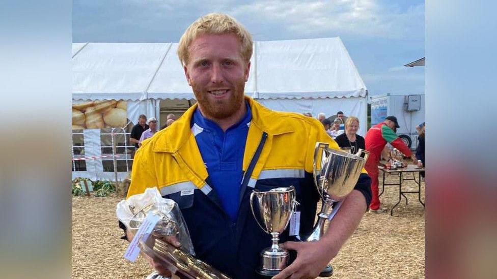 A man with blond hair and a beard holds two silver trophies in one arm and a trophy plaque in the other. He smiles at the camera while wearing a yellow and blue zip-up over a blue shirt. He is standing outside, with a white marquee in the background, and several people are outside the tent in the background.