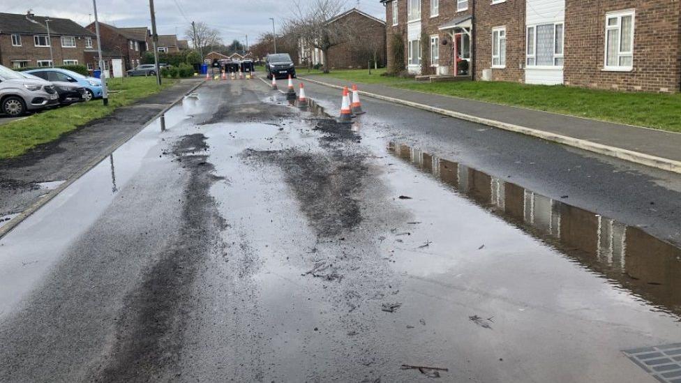 Puddles on the road with traffic cones surrounding them. There are cars parked on the left-hand side and in the distance. There are houses or flats on both sides of the road.