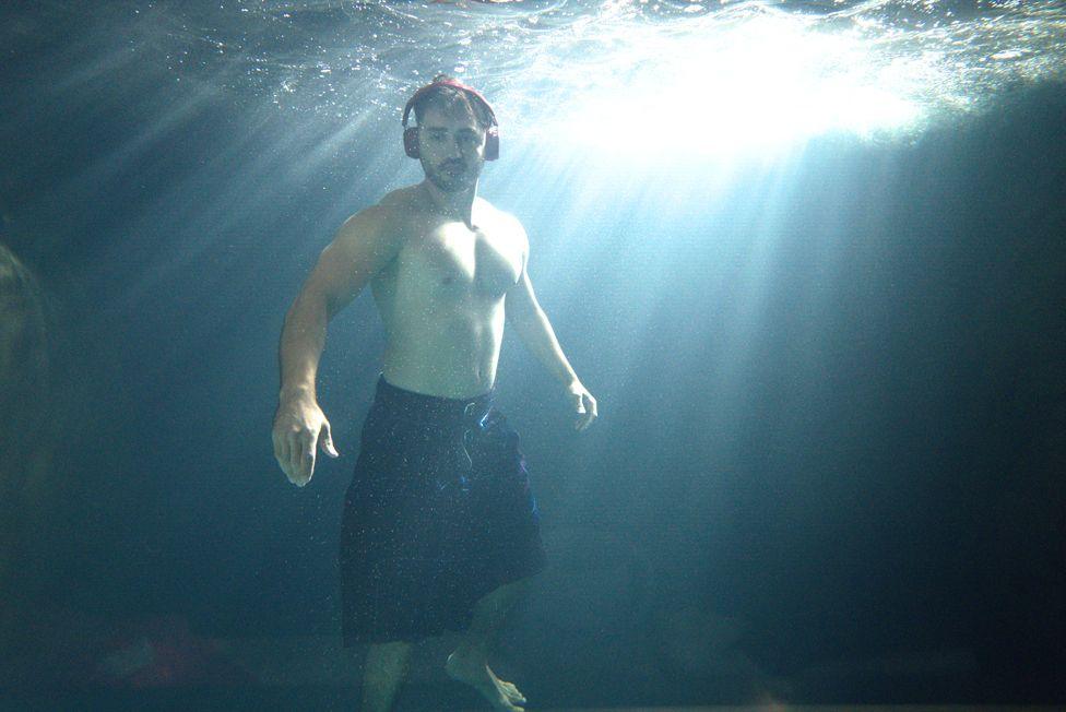 A man in a kilt with headphones on underwater with light bursting through from above