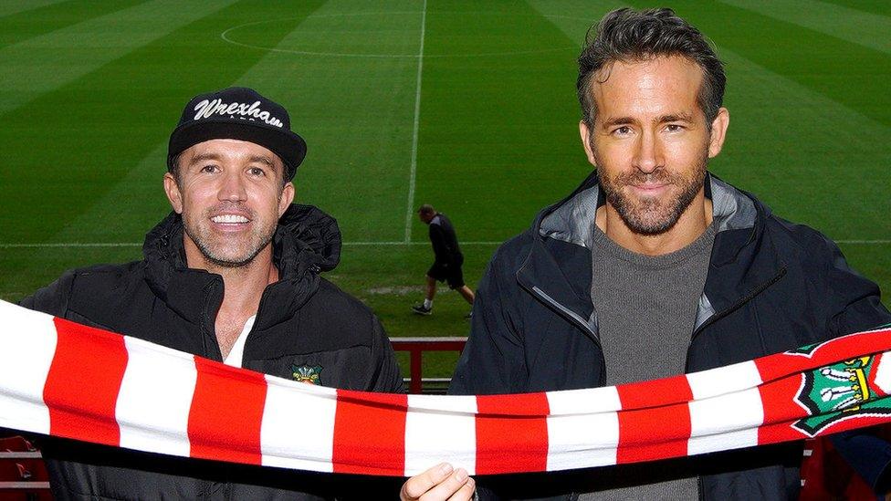 Wrexham FC owners Rob McElhenney and Ryan Reynolds smile at the camera as they hold the football club's red and white striped scarf, with the football pitch behind them.