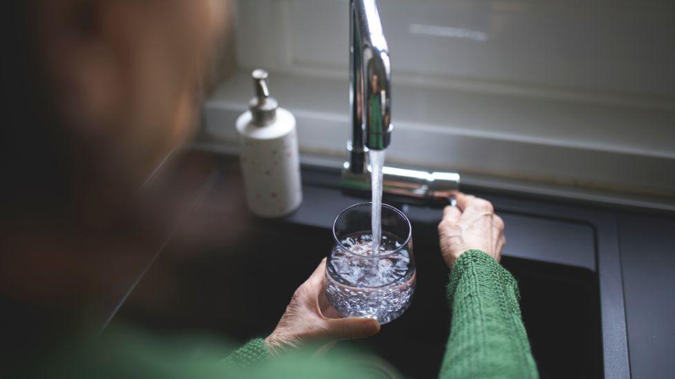 A woman using a tap to fill a glass of water. She has a green jumper on and the photo is taken over her shoulder. 