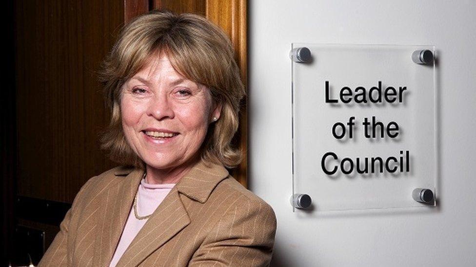 A woman leans against a doorway inside a building. She is wearing a brown suit jacket and a pink top. Next to her on the wall, there is a sign saying: "Leader of the Council."