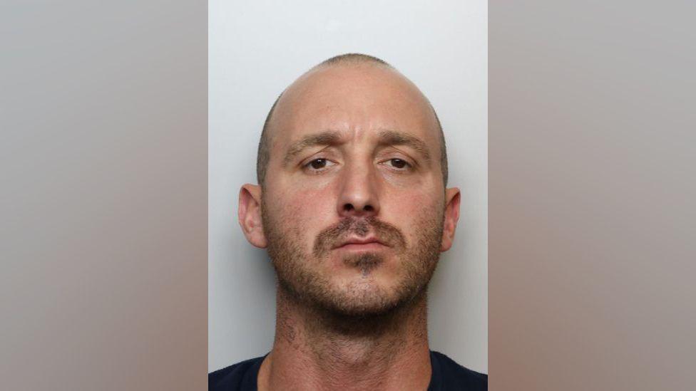 A police mugshot of Sean Deering - a man in his 30s with his shaved hair and stubble. He has brown eyes and is looking into the camera.