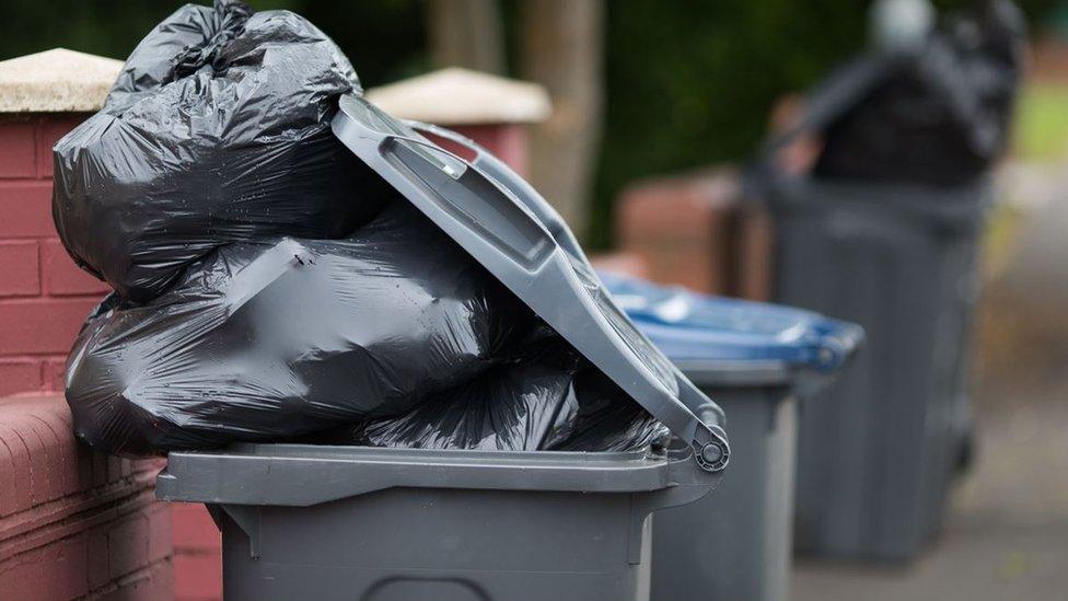 A black bin overflowing with bin bags. You can see other black bins overflowing in the background on the street