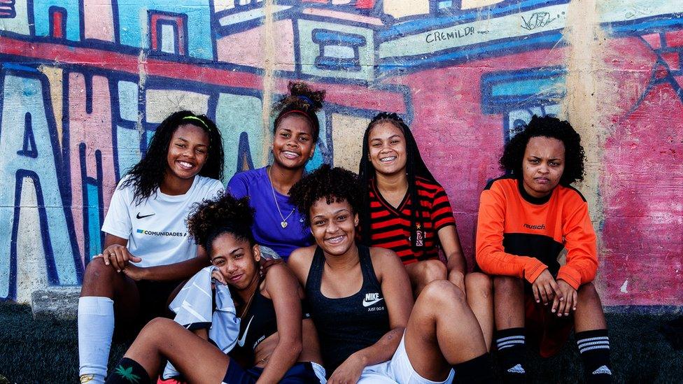 Six girls pose for the camera against the graffiti-decorated wall of their training pitch