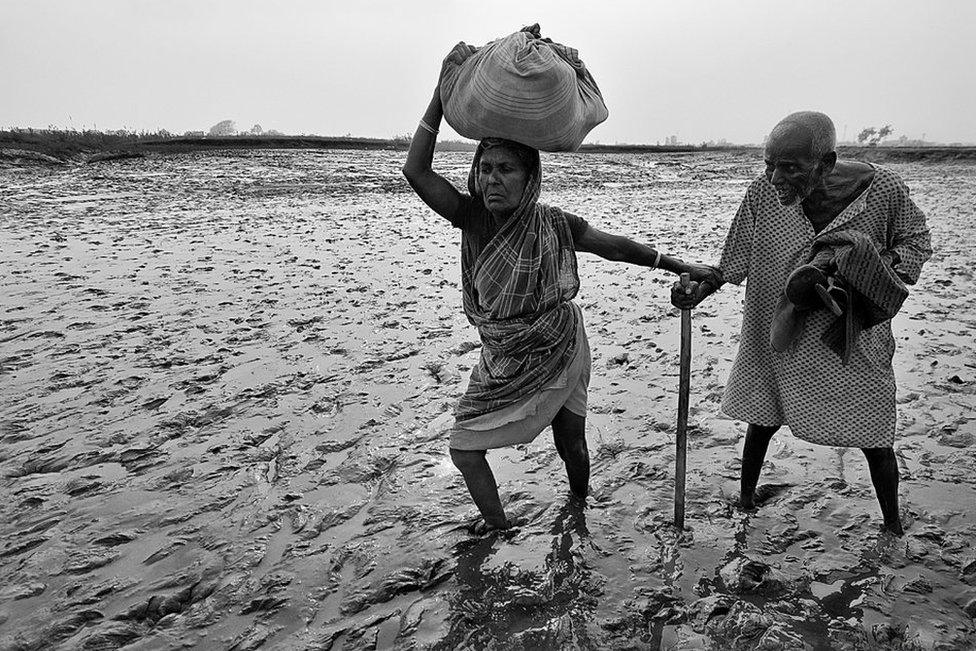 A woman helping a man through a muddy river bed at low tide