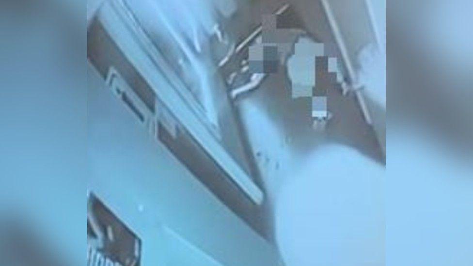 A CCTV image, with a blue-hue, showing a blurred out image of a person laying on the floor of a hallway outside a toilet door.  