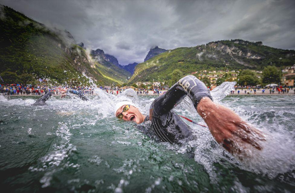 Henry Bramwell-Reeks in a wetsuit swims through choppy water during a race, with droplets splashing around. Behind the swimmer, a crowd lines the shore, and green mountains rise under a cloudy sky.