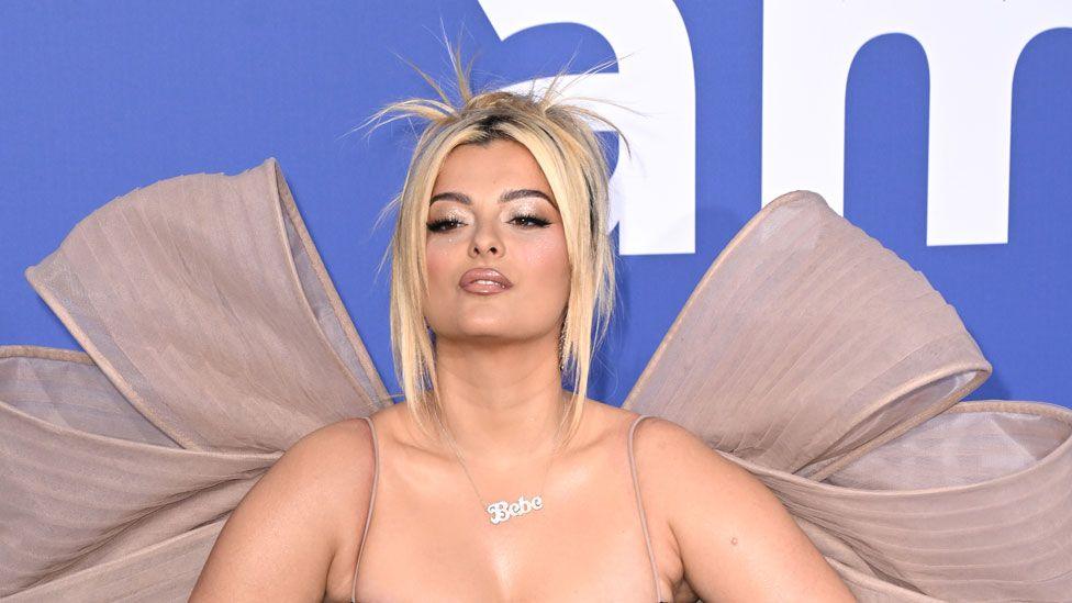 Bebe Rexha during the 76th Cannes Film Festival in Cannes, France, n 2023. She has blonde hair pulled back and around her face. She is wearing a spaghetti strap dress with a very large pink bow at her back. Her head is tilted up and she is wearing a silver necklace saying Bebe