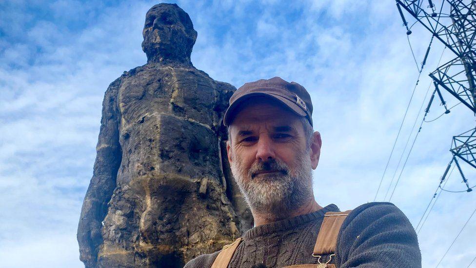 Laurence Edwards with short grey hair and beard wearing a brown cap, brown braces and a blue sweater.  The Yox Man, a huge standing bronze figure is behind him.  There is a electricity pylon to the right.