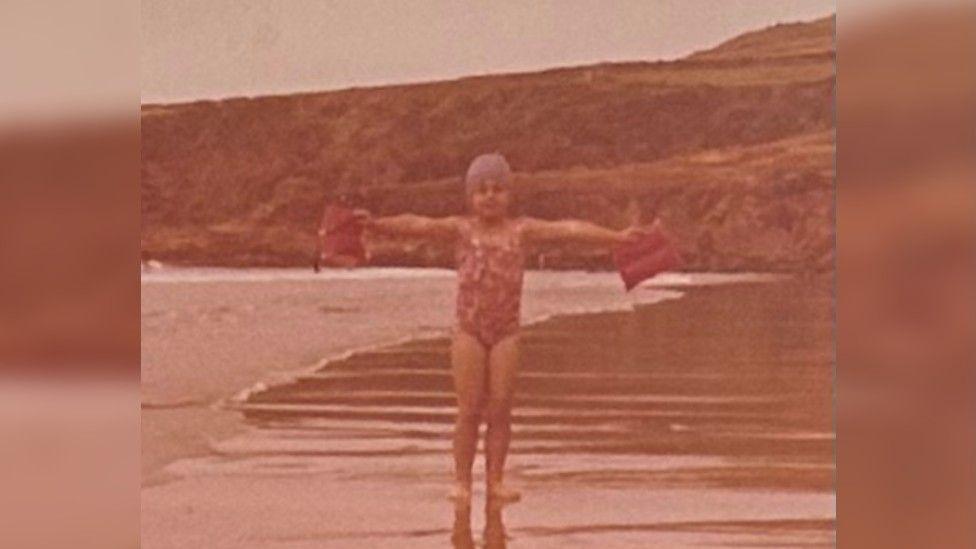 Dr Thirlwell as a young girl on a beach in a floral swimming costume and bathing hat. She is standing with her her arms raised out to the side, as if in a T shape, holding arm bands.
The picture is an old style and looks slightly aged with the sea just coming in behind her and rocky coast behind her.