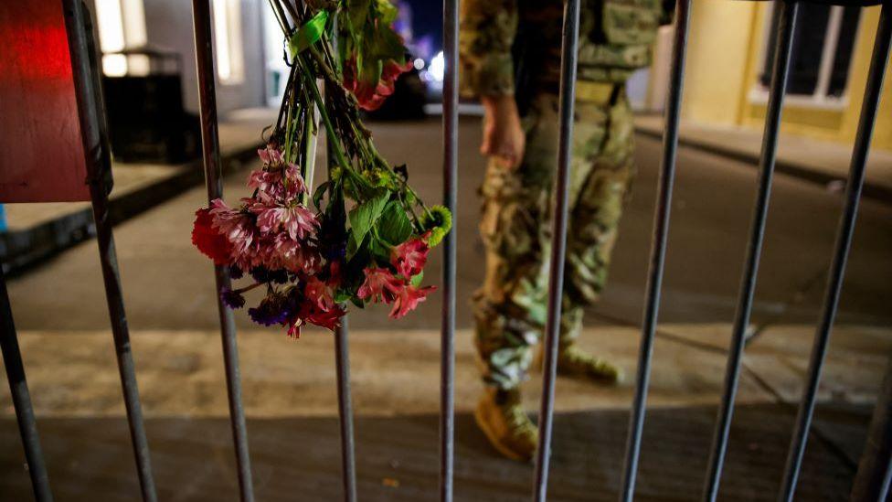 A military personnel stands near flowers on a fence near the scene