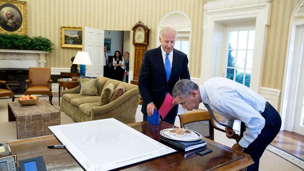 Mr Obama blows out candles as Joe Biden stands over him in the White House. Some West Wing staff members can be seen in the distant doorway, and a large framed piece of text titled "all the president's notions" lies on the desk.