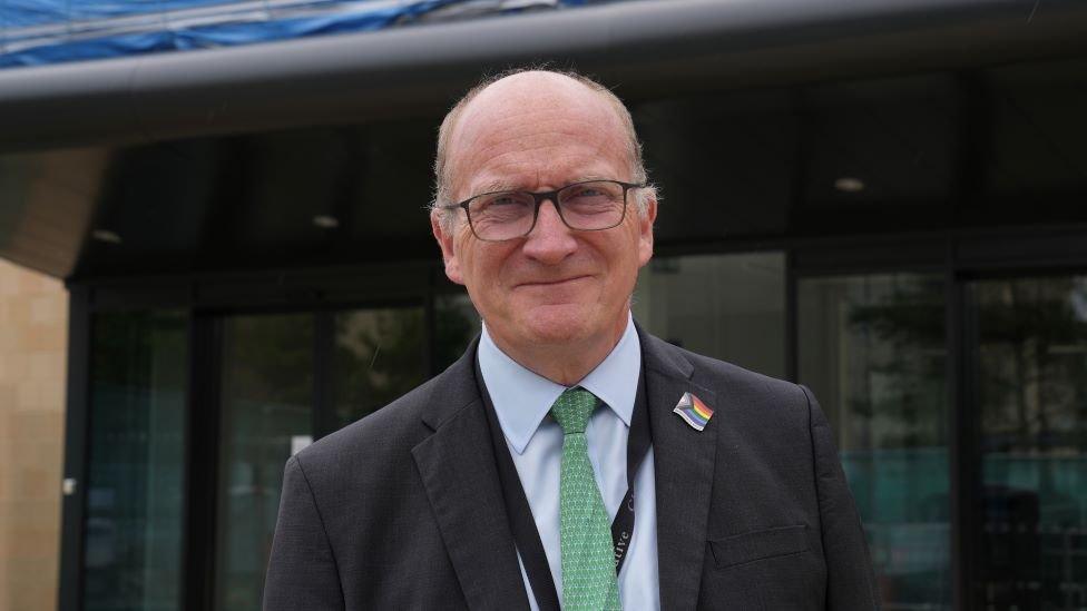 Nick Hulme stands in front of a hospital. He is wearing a light blue shirt, green tie and black suit.