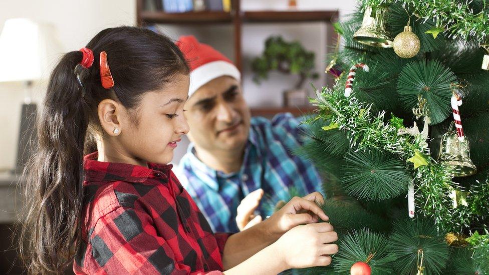 Image of a young South Asian girl putting decorations on a Christmas tree with her father in the background